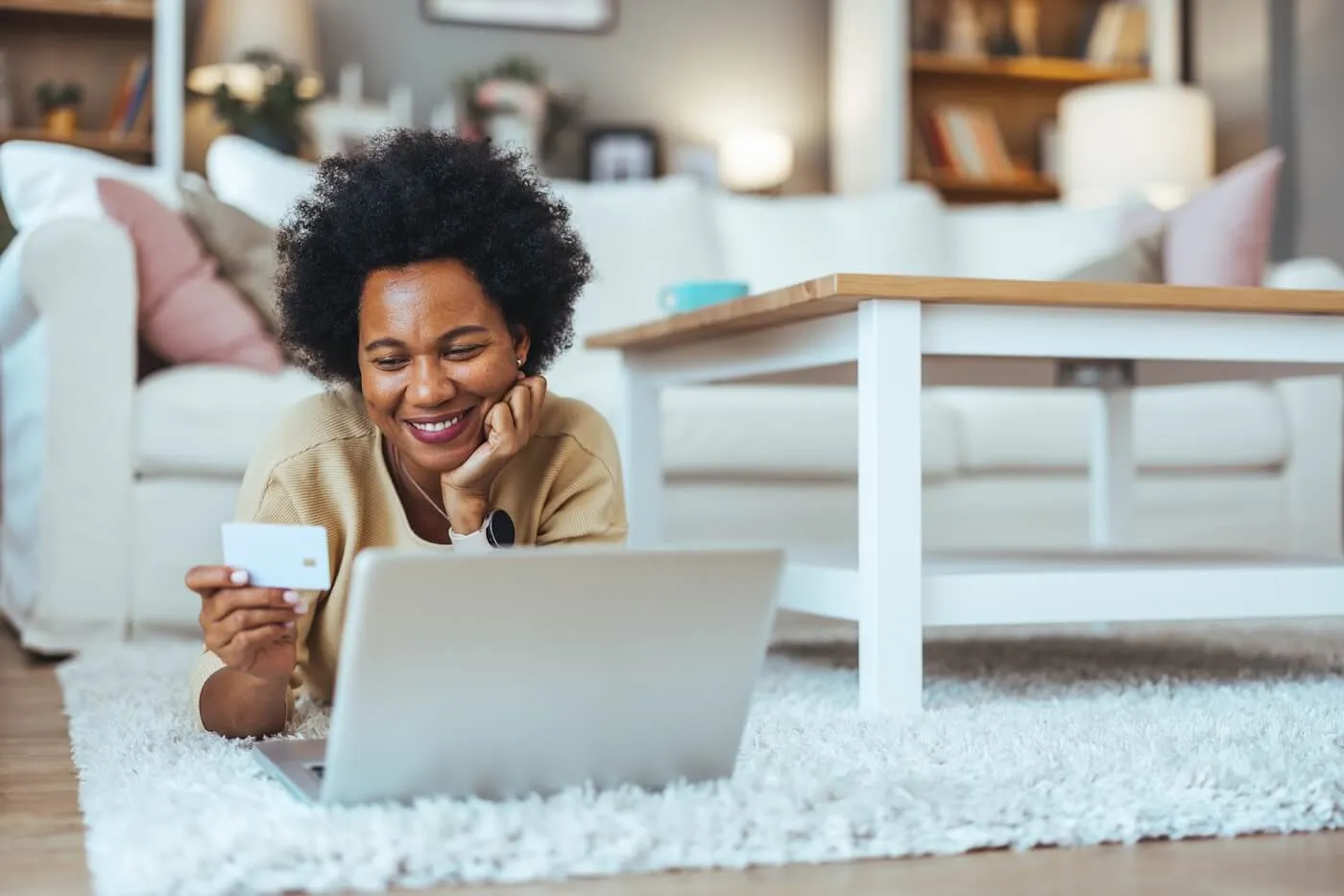 Smiling woman lying on the floor in her living room with a laptop and holding a credit card